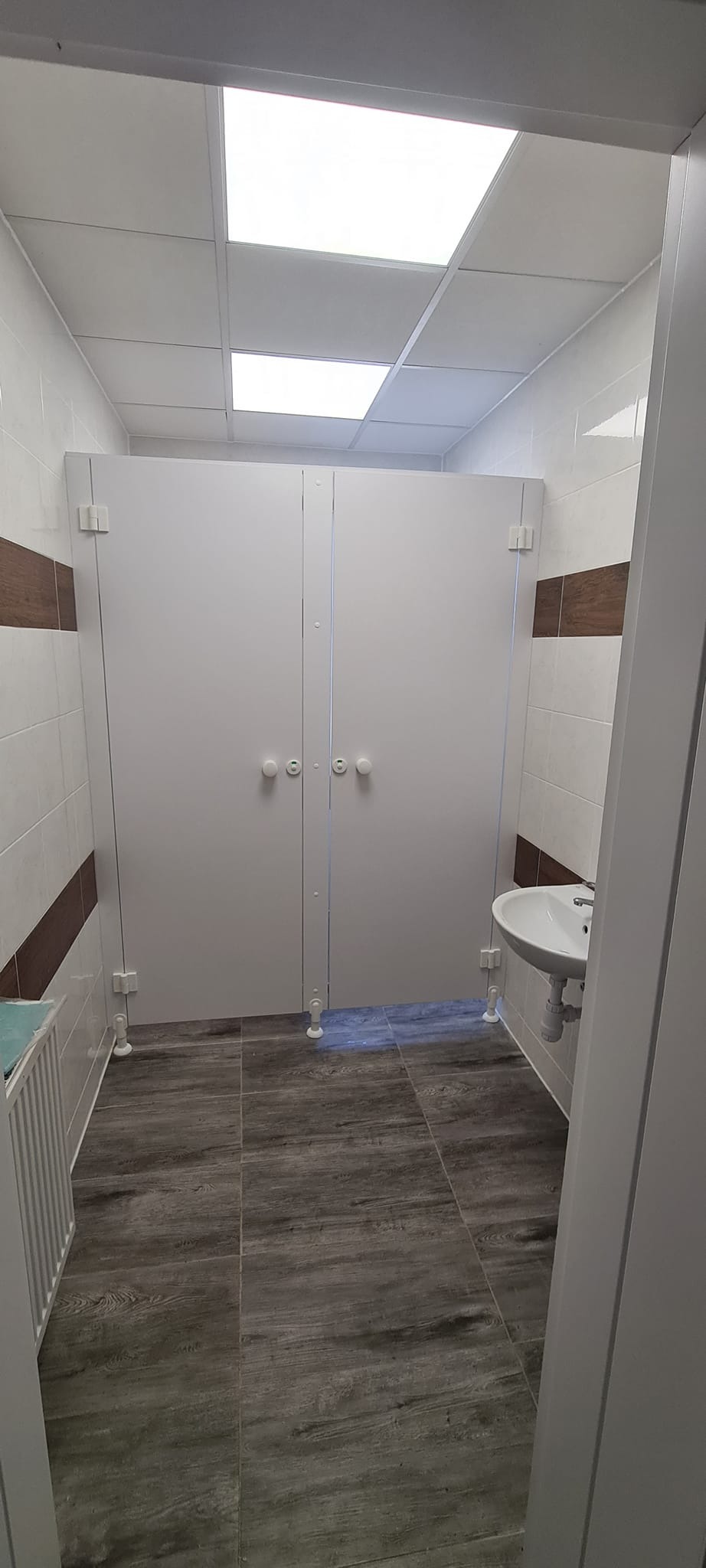 Wnętrze publicznej toalety z białymi kabinami z okrągłymi uchwytami, umywalką i podłogą imitującą drewno, oświetlone panelami sufitowymi.