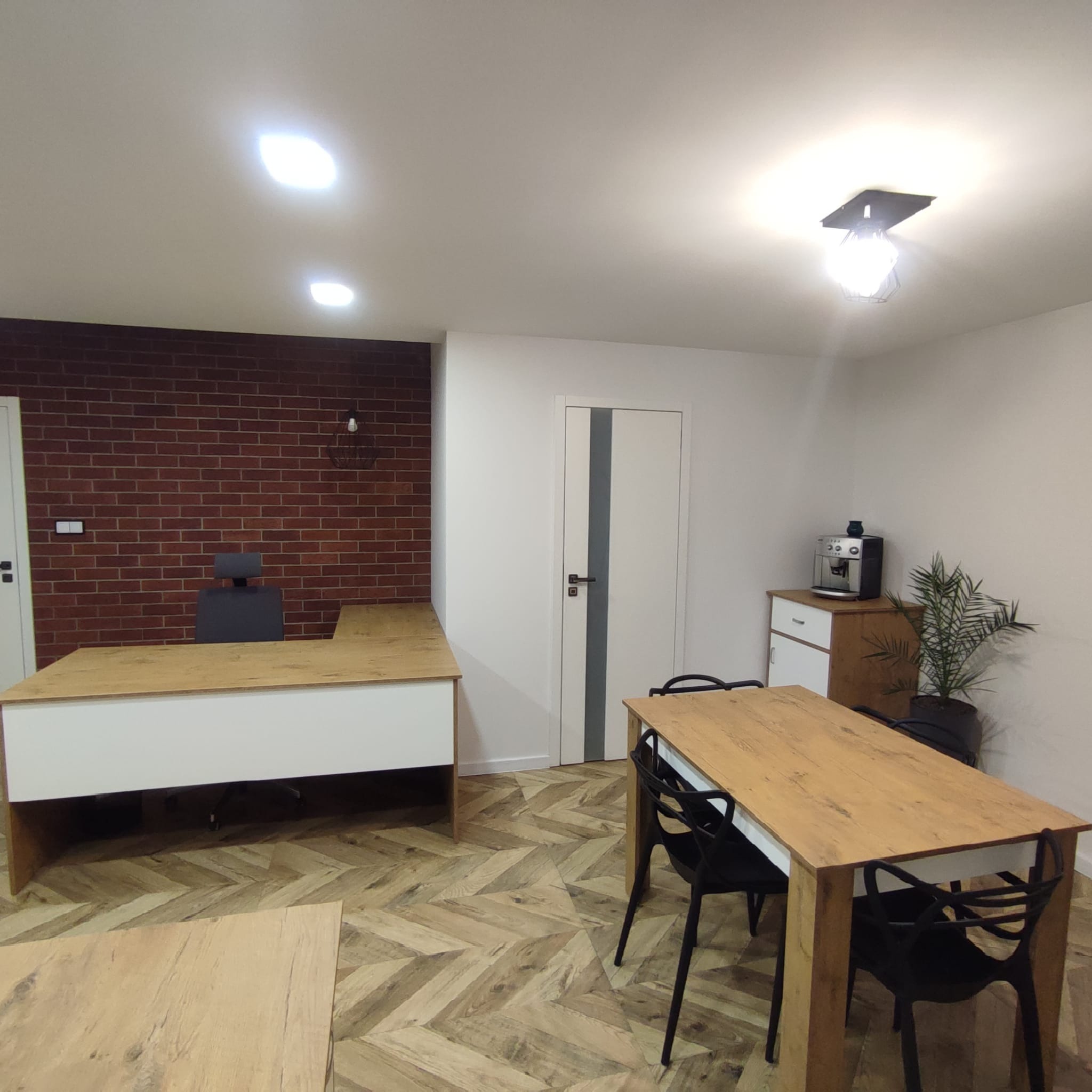 Jasne biuro z drewnianym biurkiem z białym frontem, stołem jadalnym z czarnymi krzesłami i podłogą w jodełkę, na tle ściany z czerwonej cegły oraz białej ściany z drzwiami z szarą wstawką.
