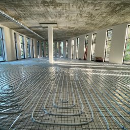 HydroAll Hydraulika - Białe rury ogrzewania podłogowego ułożone na czarnej siatce w przestronnym, jasnym pomieszczeniu z dużymi oknami i surowym betonowym sufitem.