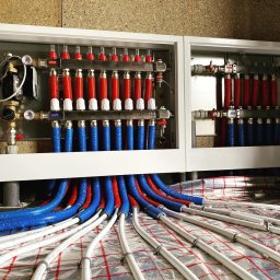 HydroAll Hydraulika - Rozdzielacz ogrzewania podłogowego w białej obudowie, z widocznymi rurami w izolacji czerwonej i niebieskiej, ułożonymi na folii grzewczej z siatką.