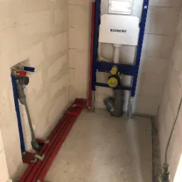 Surowy stan łazienki z zamontowanym stelażem podtynkowym Geberit, widoczne rury instalacji wodnej i kanalizacyjnej, ściany w stanie deweloperskim.