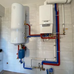 Biały piec gazowy kondensacyjny i zasobnik ciepłej wody z niebieskim naczyniem przeponowym, połączone rurami w kolorze czerwonym i niebieskim, zamontowane na ścianie z płytek ceramicznych.