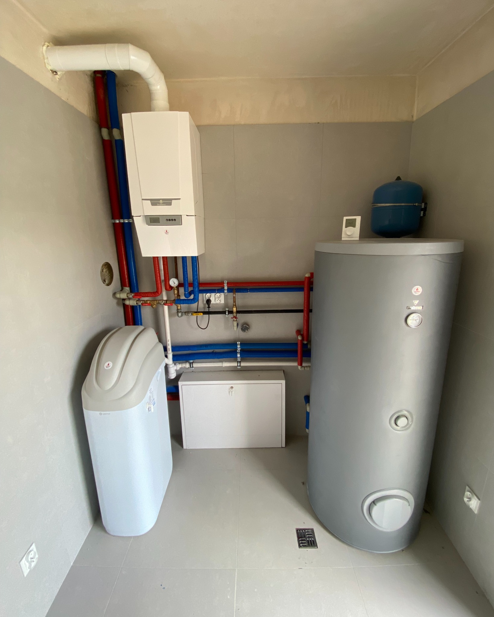 Biały piec gazowy z podłączeniami rur w kolorze niebieskim i czerwonym, obok szary zbiornik na wodę i zmiękczacz wody w pomieszczeniu z szarymi ścianami i podłogą.