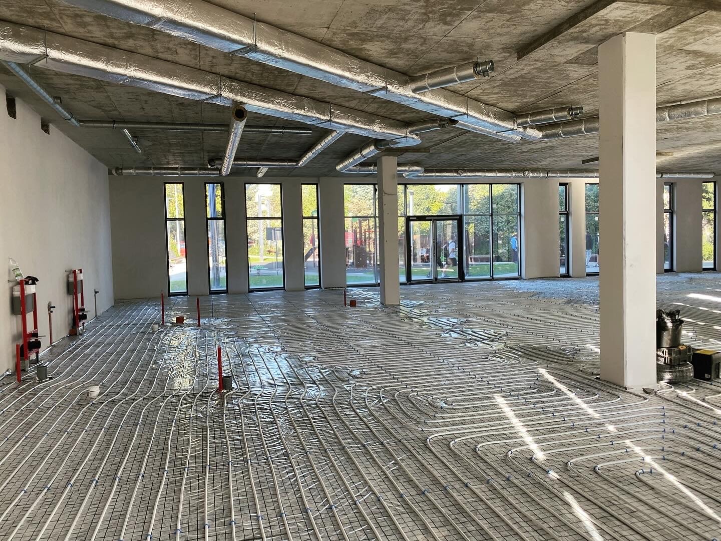 Rozległe pomieszczenie z ułożoną instalacją ogrzewania podłogowego na folii, widoczne rury wentylacyjne pod surowym betonowym sufitem i duże okna w tle.