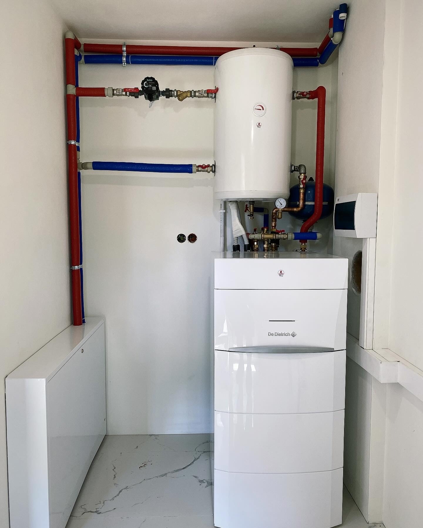 Biały piec De Dietrich z instalacją CO i CWU w pomieszczeniu z marmurową podłogą. Widoczne rury w kolorze czerwonym i niebieskim.