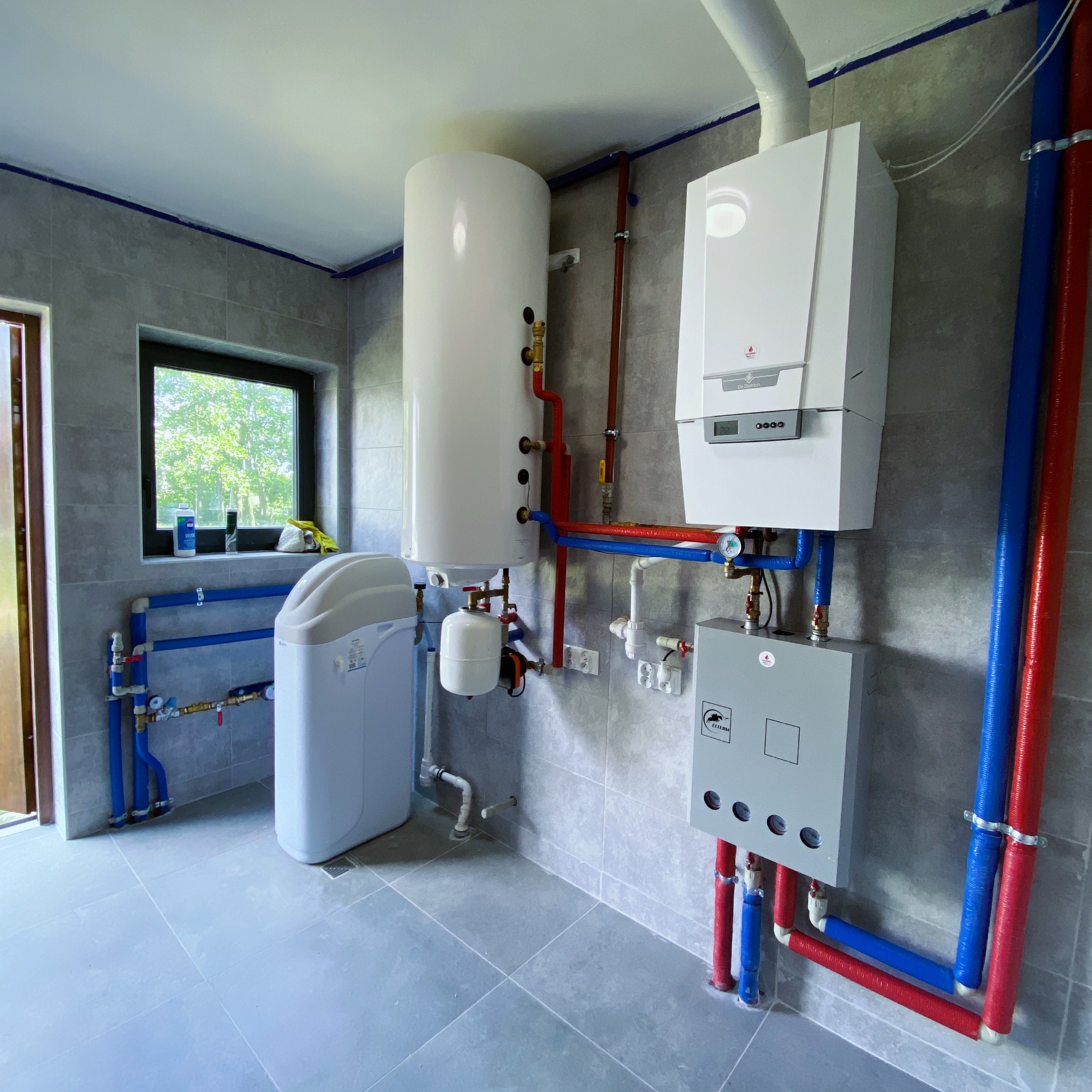 Nowoczesna kotłownia z białym zasobnikiem CWU, piecem gazowym i szarą armaturą. Widoczne rury w izolacji czerwonej i niebieskiej. Szare płytki na ścianach i podłodze.