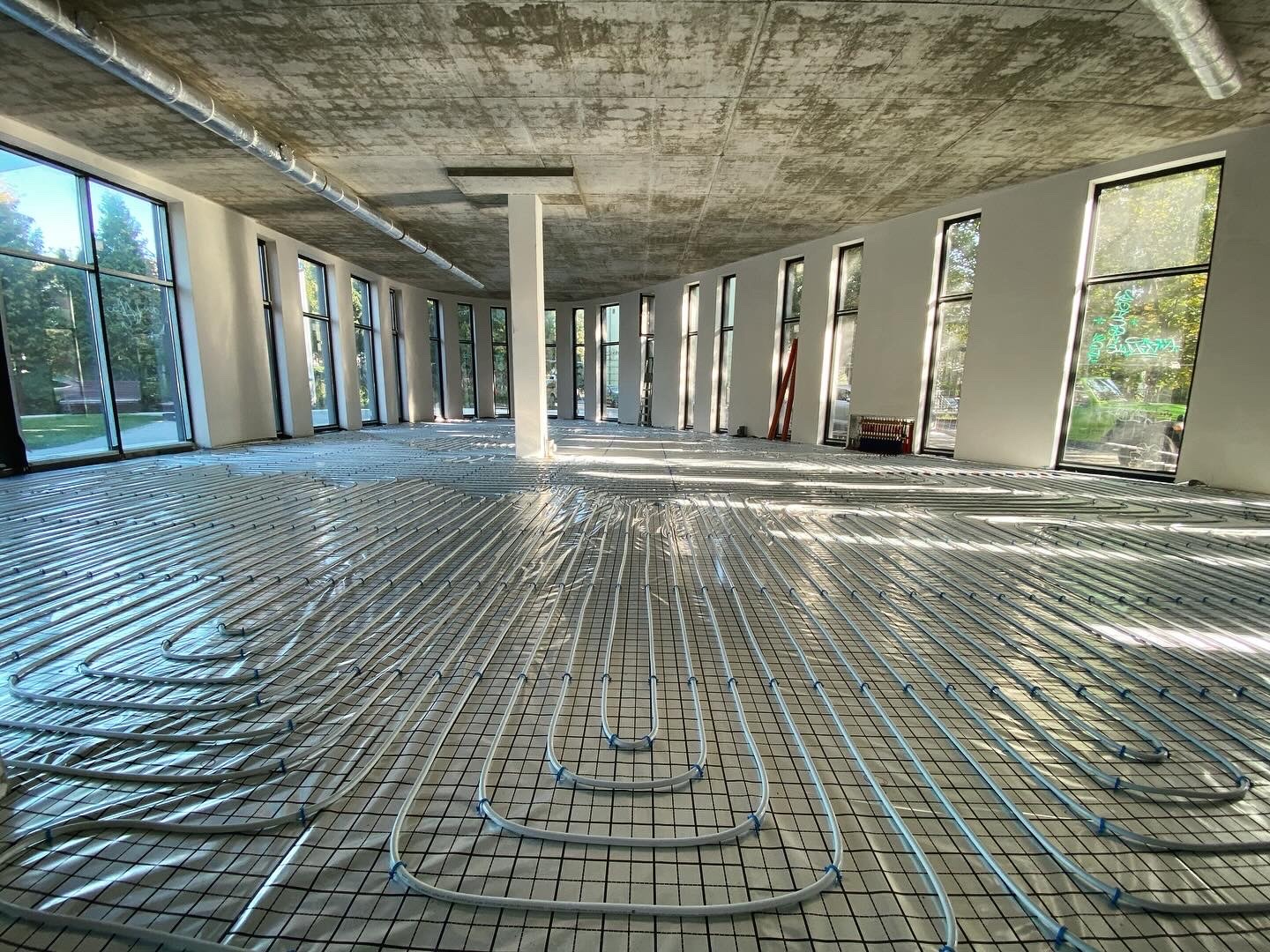 Białe rury ogrzewania podłogowego ułożone na czarnej siatce w przestronnym, jasnym pomieszczeniu z dużymi oknami i surowym betonowym sufitem.