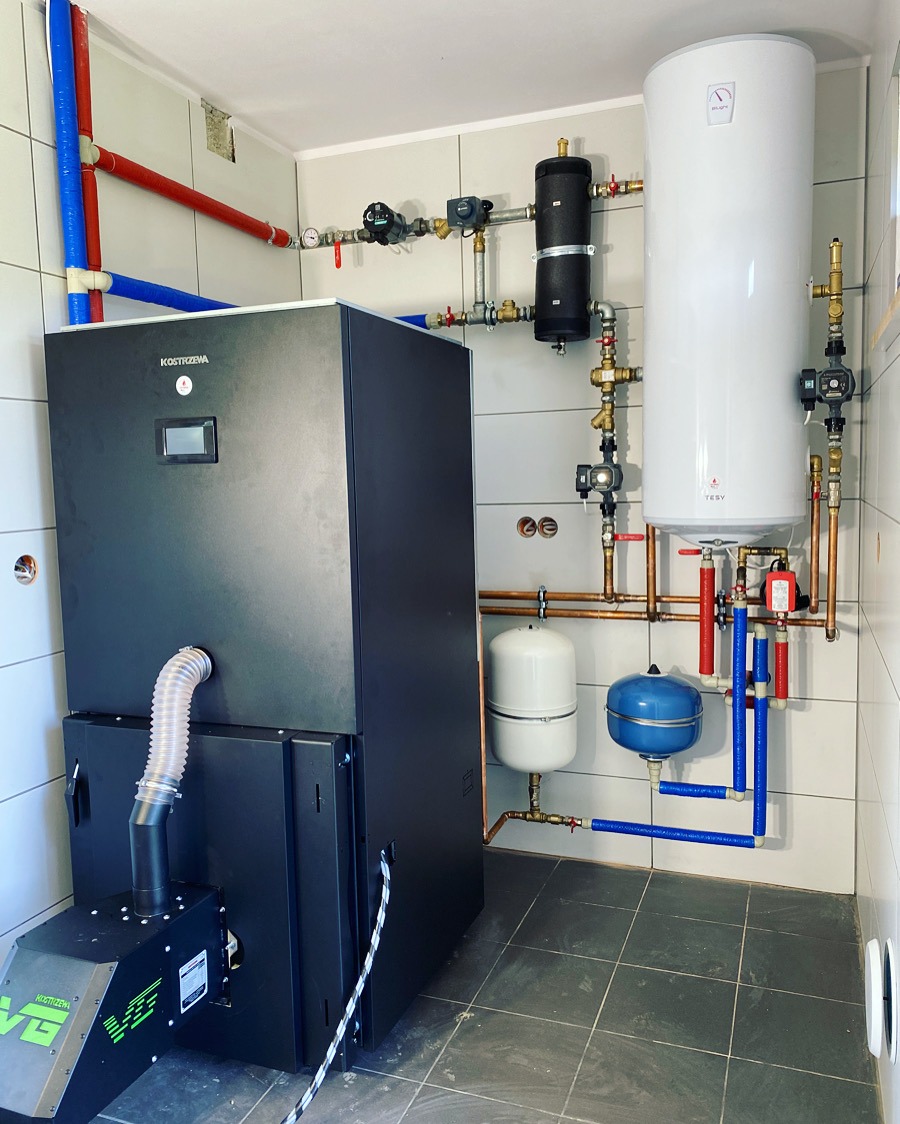 Nowoczesna kotłownia z piecem Kostrzewa, zasobnikiem ciepłej wody i rozbudowaną instalacją hydrauliczną w pomieszczeniu wykończonym płytkami.
