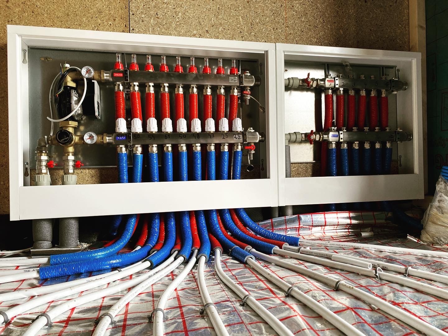 Rozdzielacz ogrzewania podłogowego w białej obudowie, z widocznymi rurami w izolacji czerwonej i niebieskiej, ułożonymi na folii grzewczej z siatką.