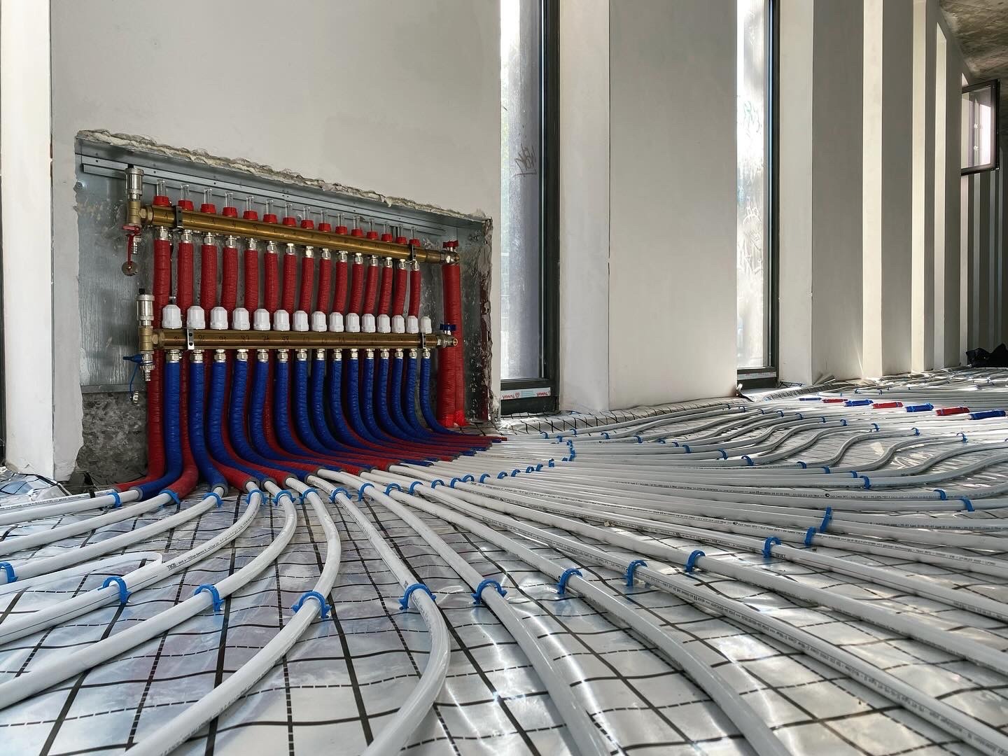 Rozłożona instalacja ogrzewania podłogowego z rozdzielaczem na ścianie, rury w kolorach czerwonym i niebieskim. Widoczne okna w tle, folia izolacyjna pod rurami.
