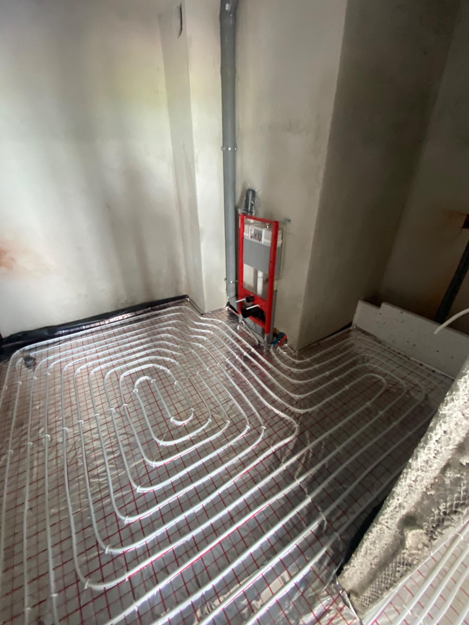 Rozłożone rury ogrzewania podłogowego na folii z siatką, widoczny pion kanalizacyjny i stelaż podtynkowy WC w surowym stanie.