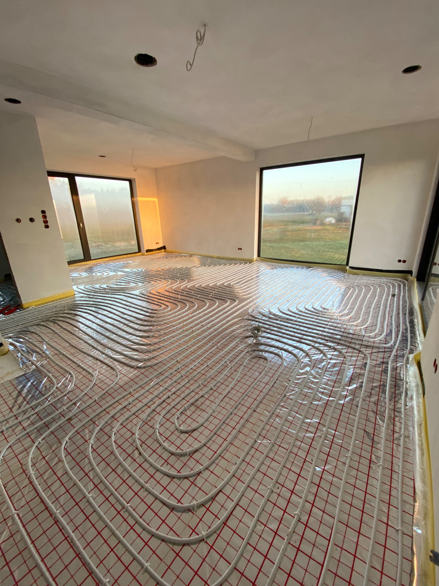 Białe rurki ogrzewania podłogowego ułożone na czerwonej siatce w przestronnym, jasnym pomieszczeniu z dużymi oknami, zabezpieczone folią i taśmą malarską.
