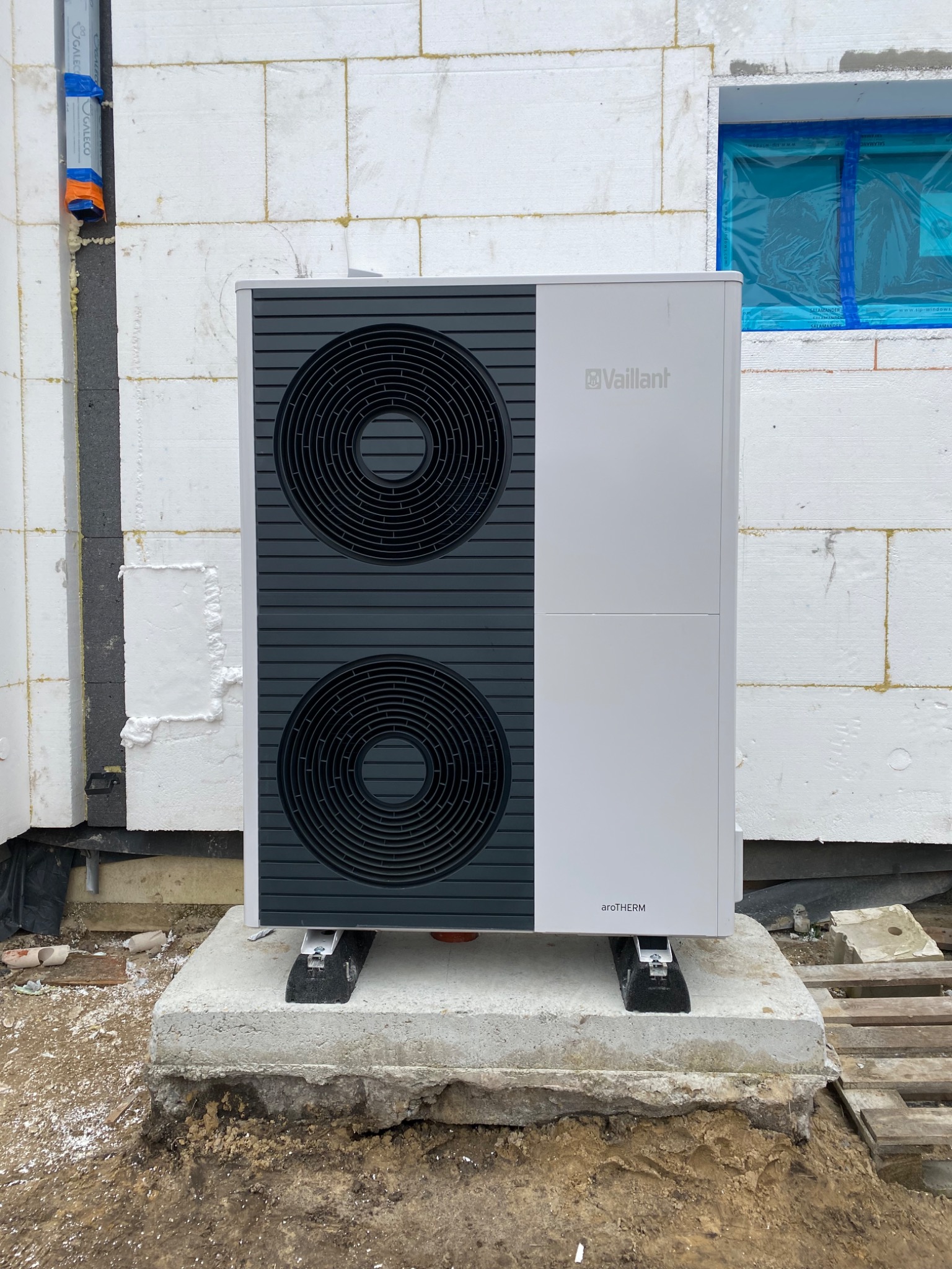 Nowoczesna pompa ciepła Vaillant aroTHERM zamontowana na betonowym fundamencie przed budynkiem w trakcie budowy, widoczne białe bloczki i okno zabezpieczone folią.