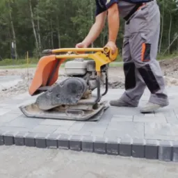 Pracownik w szarych spodniach roboczych zagęszcza świeżo ułożoną, szarą kostkę brukową za pomocą pomarańczowo-żółtej zagęszczarki spalinowej, widoczne obrzeża z ciemnej kostki.