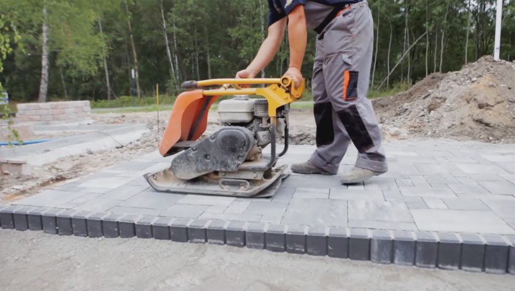Pracownik w szarych spodniach roboczych zagęszcza świeżo ułożoną, szarą kostkę brukową za pomocą pomarańczowo-żółtej zagęszczarki spalinowej, widoczne obrzeża z ciemnej kostki.