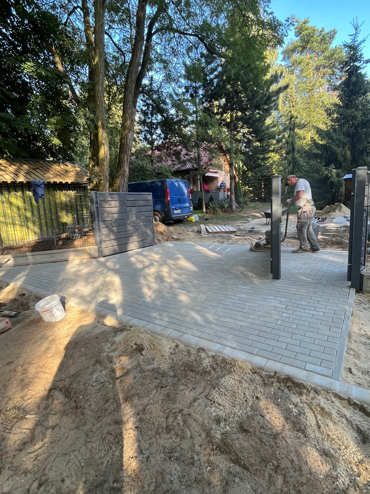 Montaż bramy wjazdowej na posesję z częściowo ułożoną kostką brukową, widoczny pracownik z zagęszczarką, brama w kolorze grafitowym, w tle budynek mieszkalny i zaparkowany bus.