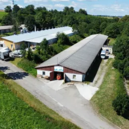 Widok z lotu ptaka na budynek magazynowy z otwartymi wrotami, przez które wyjeżdża pomarańczowy wózek widłowy z paletami. W tle inne budynki i zalesiony teren.