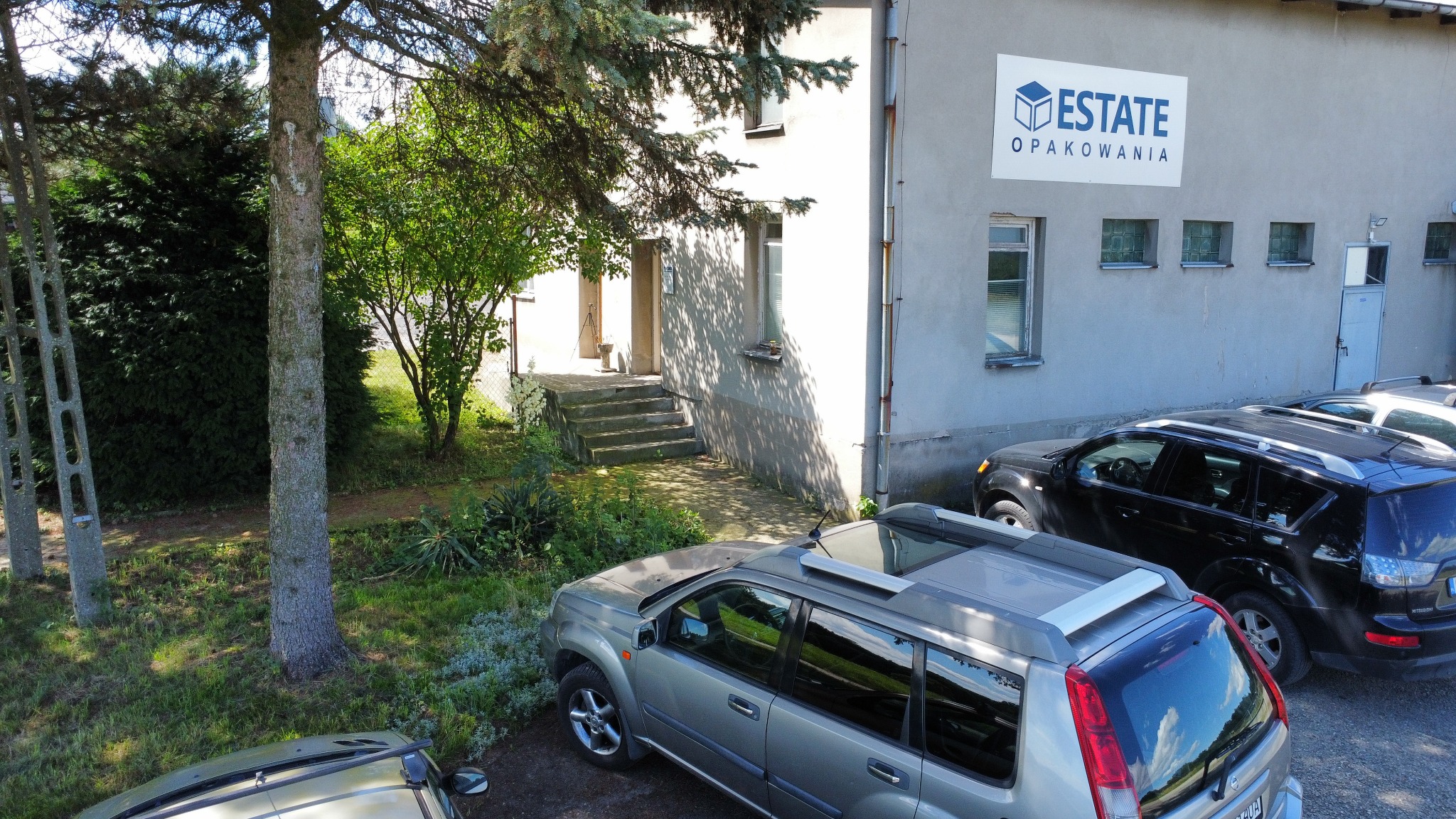 Budynek firmy z logo 'Estate Opakowania' na ścianie, widok z góry na zaparkowane samochody, drzewa i wejście do budynku.