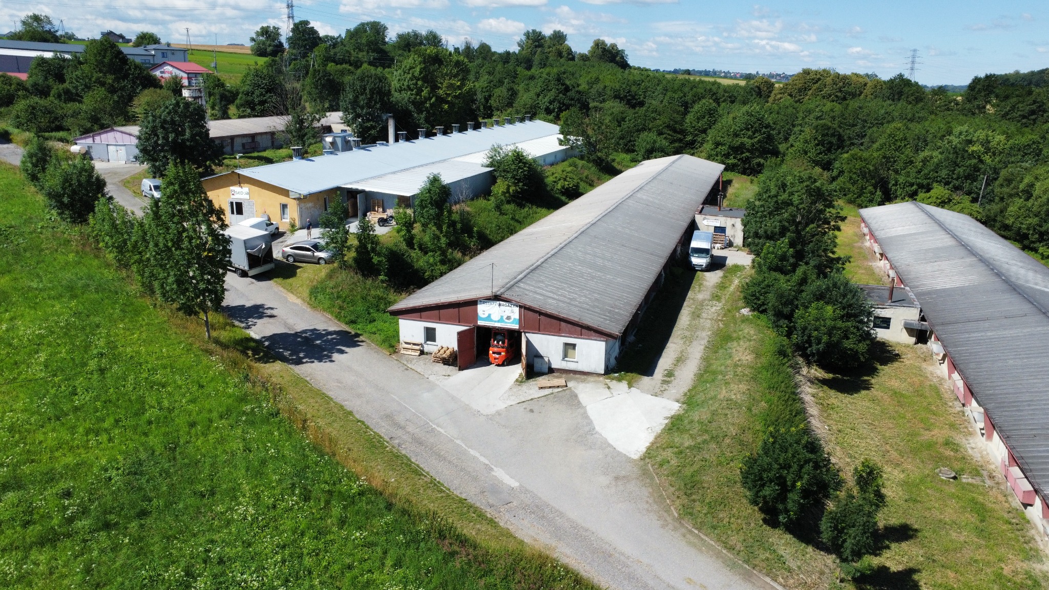 Widok z lotu ptaka na budynek magazynowy z otwartymi wrotami, przez które wyjeżdża pomarańczowy wózek widłowy z paletami. W tle inne budynki i zalesiony teren.