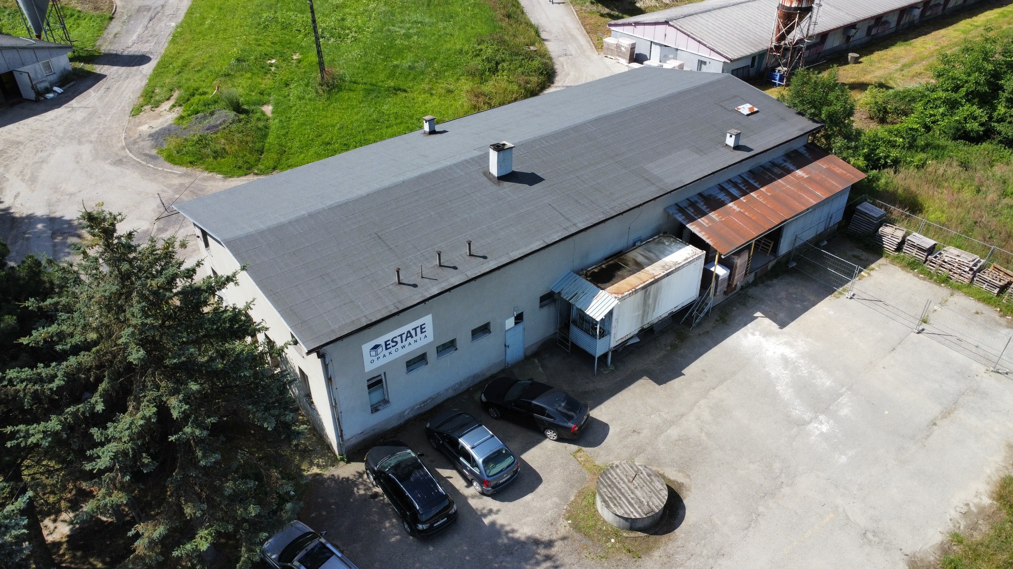 Widok z drona na budynek firmy Estate Opakowania z zaparkowanymi samochodami, zaroślami i częścią terenu przemysłowego w tle.