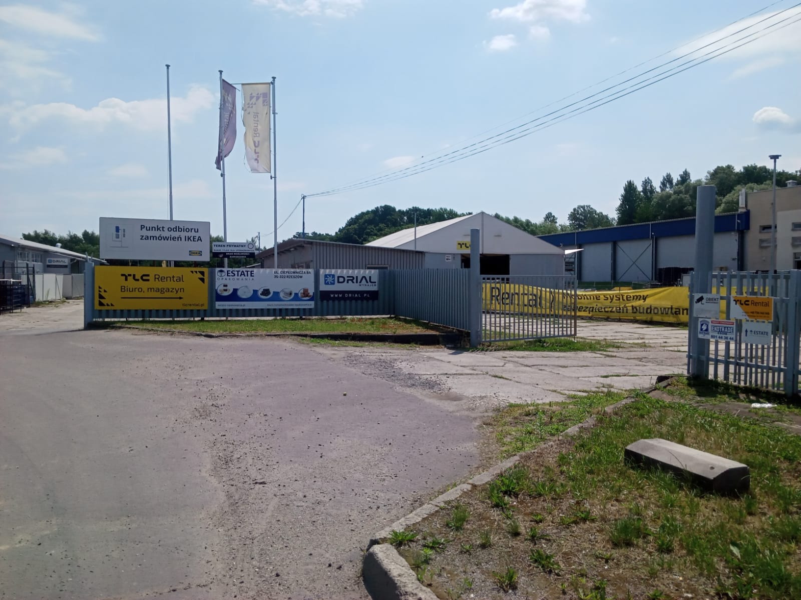 Widok na teren przemysłowy z różnymi firmami, w tym punkt odbioru zamówień IKEA, TLC Rental biuro i magazyn, Estate Opalowania oraz Drial, z widocznymi banerami reklamowymi i flagami