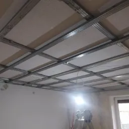 Montaż sufitu podwieszanego z płyt gipsowych na metalowej konstrukcji, widoczna drabina i przewody elektryczne.