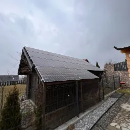 Instalacja fotowoltaiczna na dachu małego, drewnianego budynku gospodarczego, widok z poziomu gruntu, pochmurne niebo w tle.