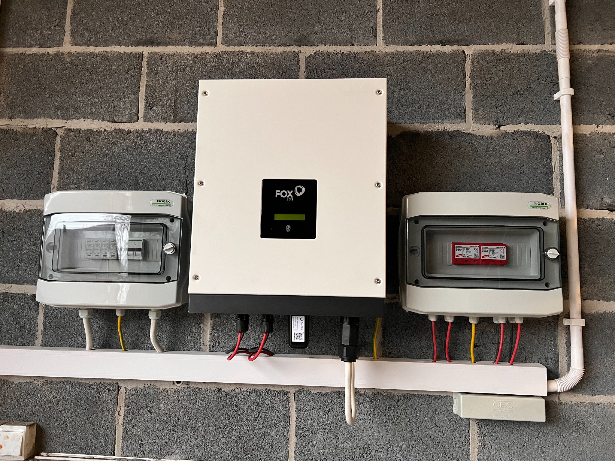 Instalacja elektryczna z inwerterem FOX ESS i zabezpieczeniami przepięciowymi Noark, zamontowana na szarej ścianie z bloczków betonowych.