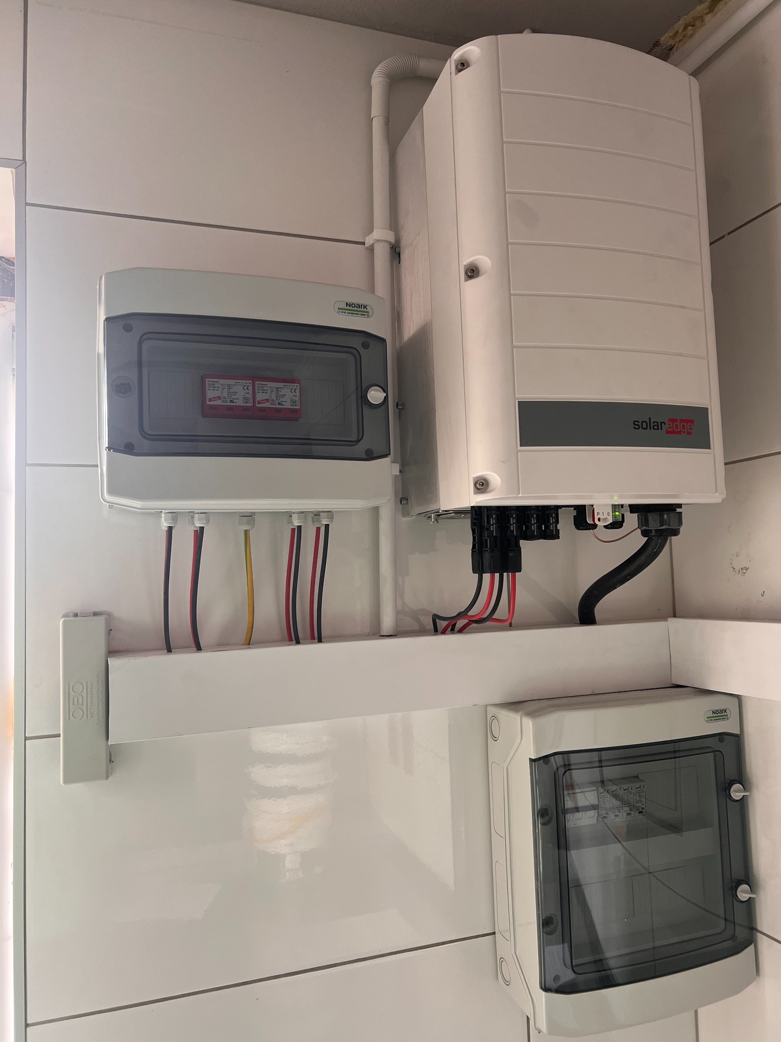 Instalacja elektryczna: biała skrzynka z bezpiecznikami podłączona przewodami w kolorach czarnym, czerwonym i żółtym, obok duży biały inwerter SolarEdge zamontowany na białej ścianie z płytek.