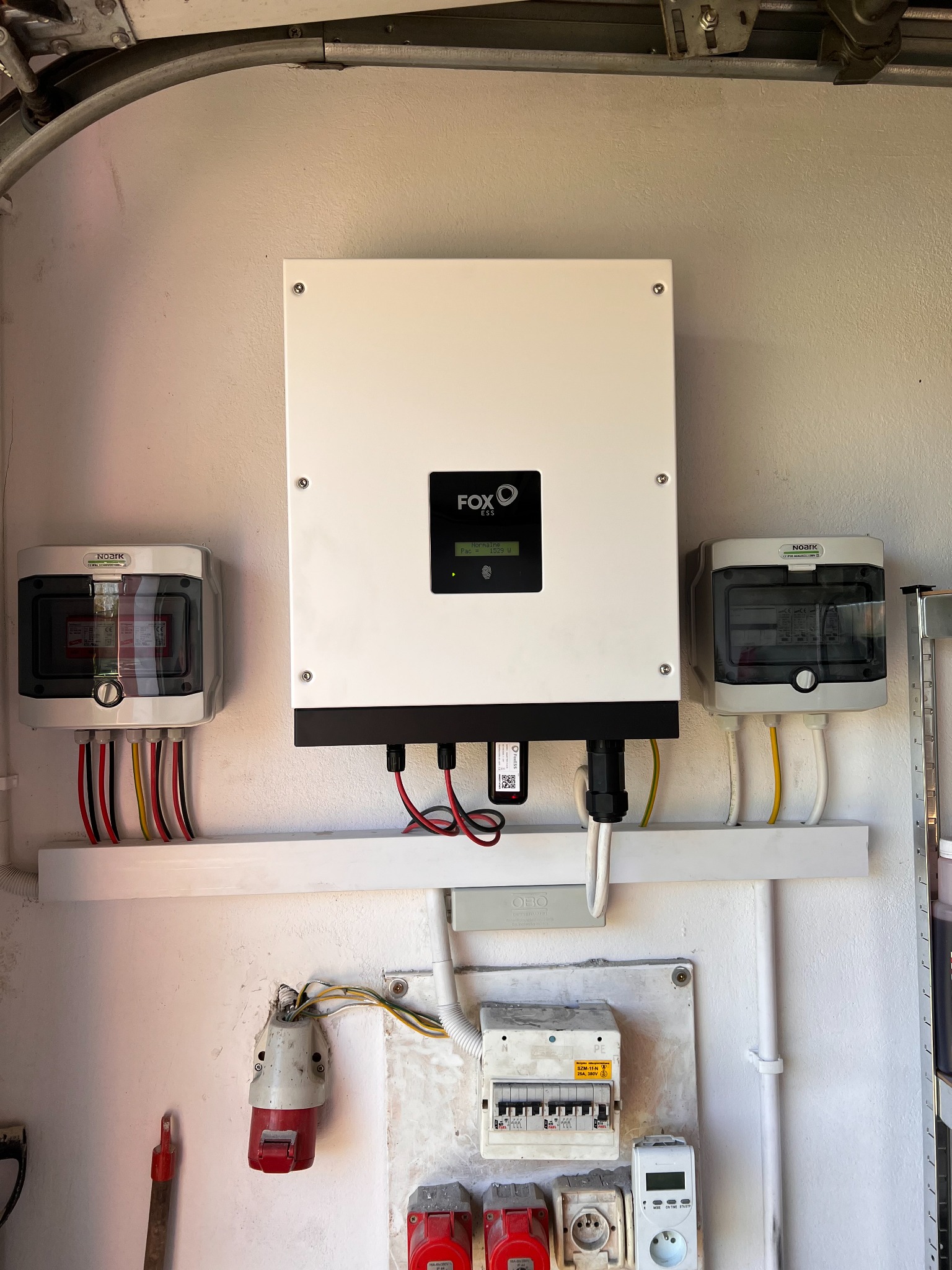 Biały falownik marki FoxESS zamontowany na ścianie w garażu, z widocznymi zabezpieczeniami Noark i okablowaniem elektrycznym. Na ścianie poniżej widoczne gniazda siłowe i bezpieczniki.
