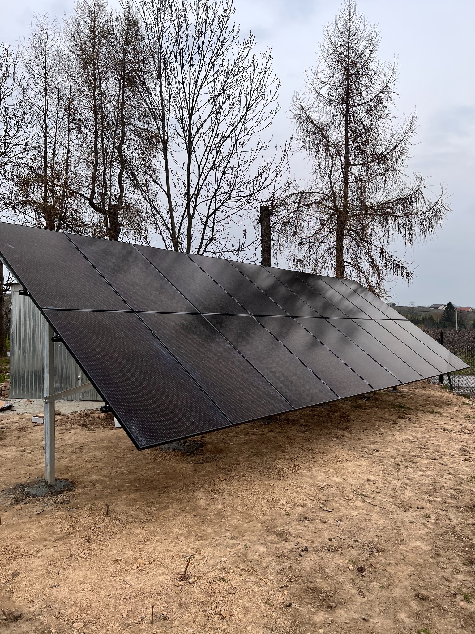 Panel fotowoltaiczny ustawiony na ziemi, widoczne metalowe wsporniki, w tle drzewa bez liści i zabudowania.