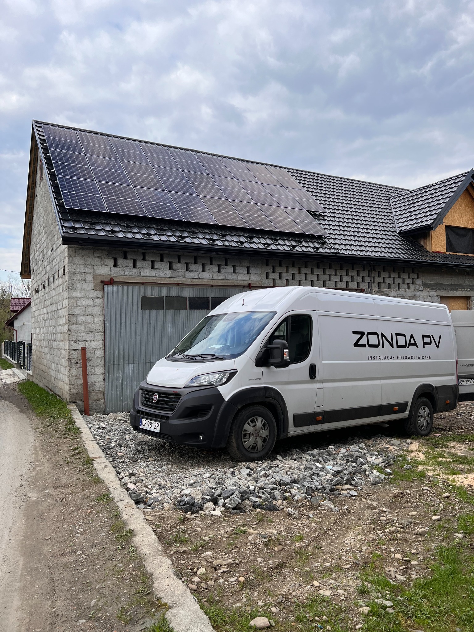 Szary budynek z zamontowanymi panelami słonecznymi na dachu, obok zaparkowany biały samochód dostawczy z napisem ZONDA PV INSTALACJE FOTOWOLTAICZNE.