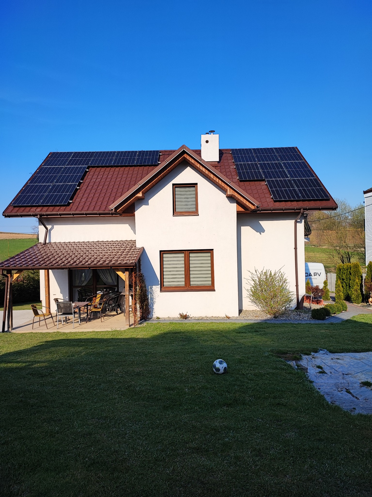 Biały dom jednorodzinny z brązowym dachem i panelami słonecznymi na dachu, widok z ogrodu z piłką do nogi na trawniku.