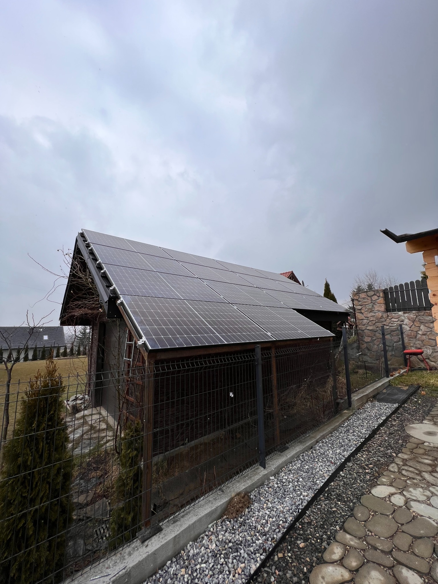 Instalacja fotowoltaiczna na dachu małego, drewnianego budynku gospodarczego, widok z poziomu gruntu, pochmurne niebo w tle.