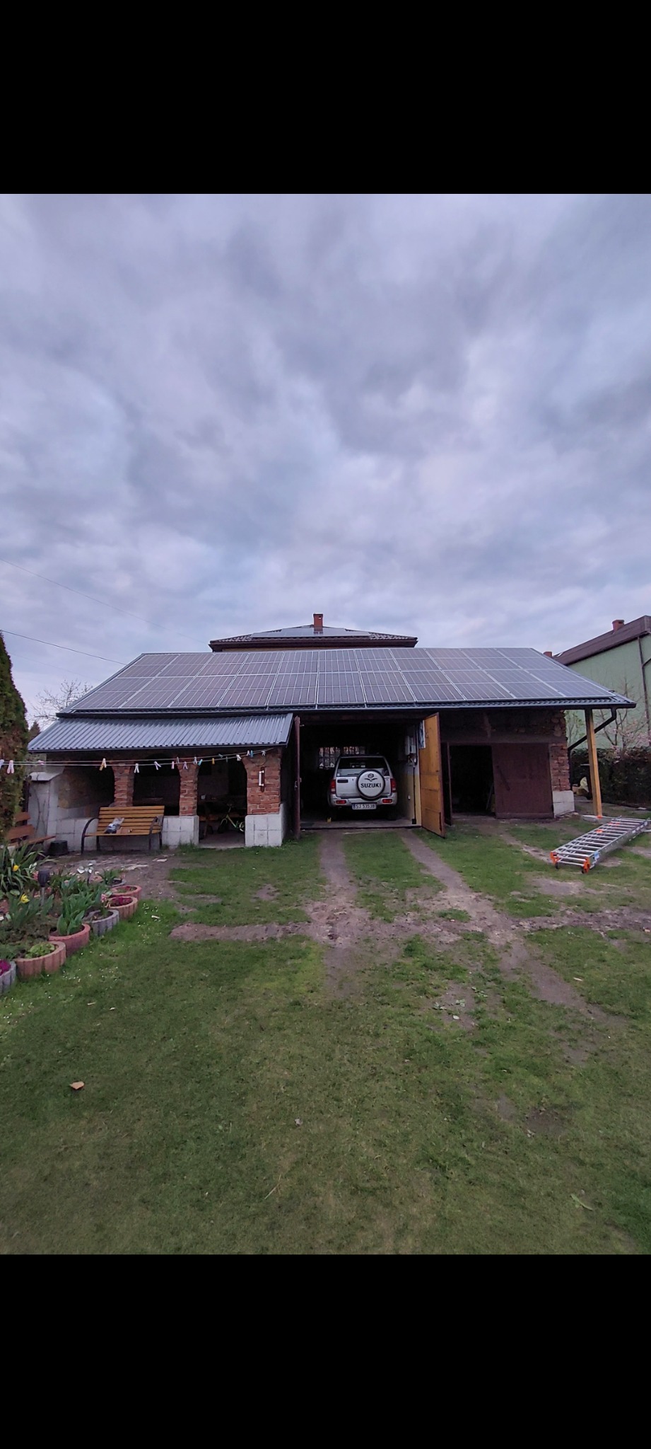 Panele fotowoltaiczne na dachu budynku gospodarczego z cegły, z otwartymi wrotami garażowymi, w tle pochmurne niebo.