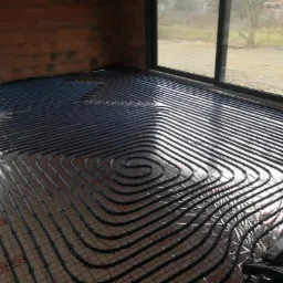 Układanie rur ogrzewania podłogowego na folii izolacyjnej w surowym stanie, widoczne okna i ceglany fragment ściany.