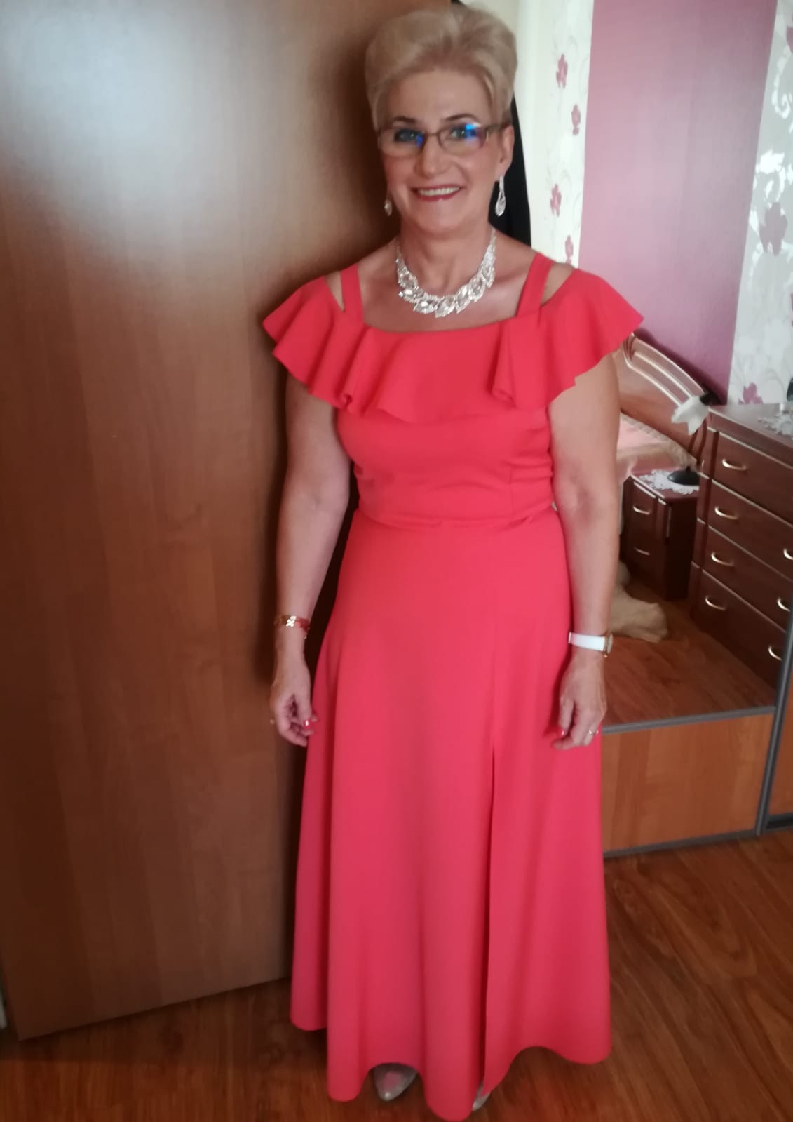 Uśmiechnięta kobieta w okularach i długiej, koralowej sukni z falbanami, pozuje w pomieszczeniu z drewnianymi meblami i jasną tapetą w kwiaty.