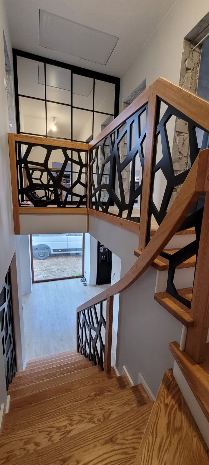 Drewniane schody z ażurową, czarną balustradą, widok z góry na dół, na dole duże okno z widokiem na samochód dostawczy.