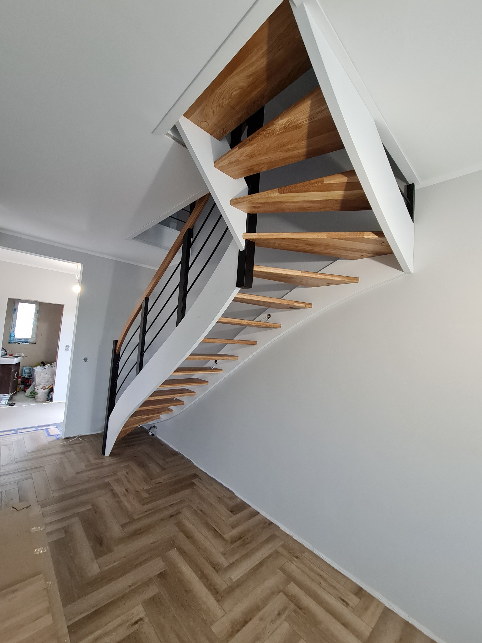 Nowoczesne schody drewniane z czarnymi metalowymi balustradami i białymi elementami konstrukcyjnymi, widok z dołu na tle jasnego wnętrza z podłogą ułożoną w jodełkę.