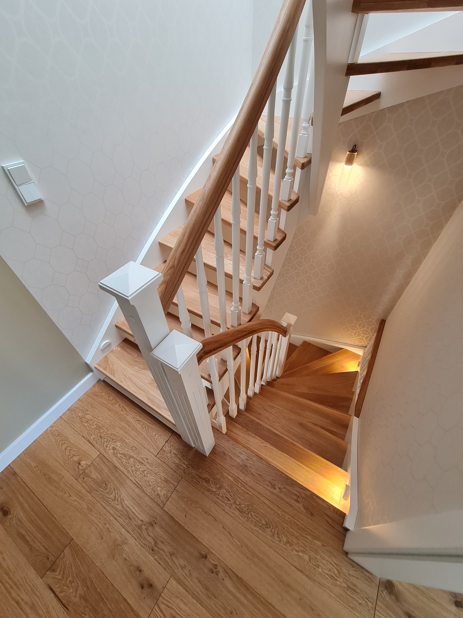 Drewniane schody z białymi balustradami, oświetlone od spodu, widok z góry na parkietową podłogę i wzorzystą tapetę.