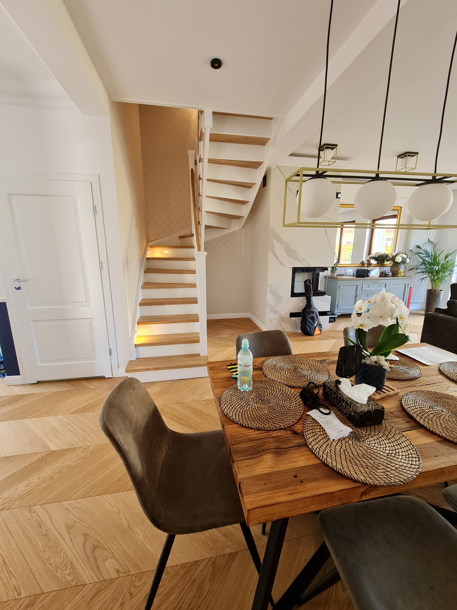 Jasne wnętrze domu z drewnianą podłogą w jodełkę, schodami z podświetleniem LED, stołem jadalnym z dekoracjami i lampą wiszącą z trzema białymi kloszami.