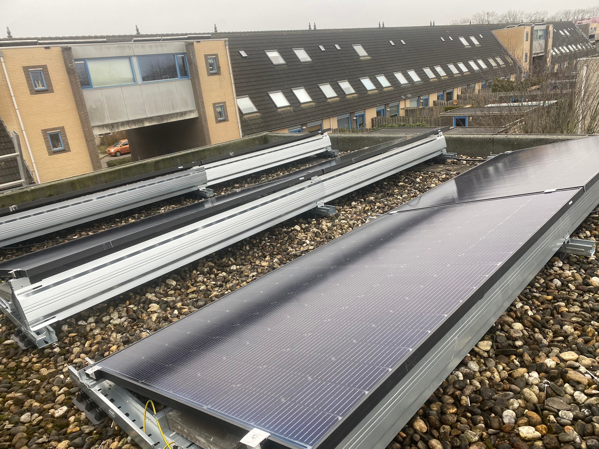 Panele słoneczne w trakcie montażu na dachu pokrytym drobnymi kamieniami; widoczne aluminiowe profile montażowe i fragmenty instalacji elektrycznej.