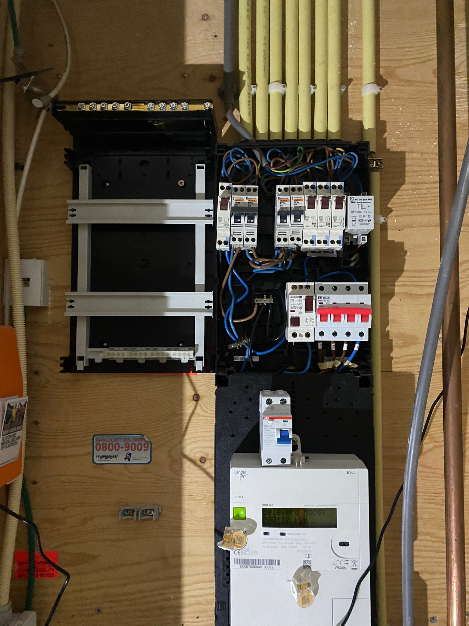 Otwarta skrzynka bezpiecznikowa z widocznymi bezpiecznikami, przewodami i licznikiem energii elektrycznej zamontowana na drewnianej ścianie. Nad skrzynką widoczne rury instalacyjne.