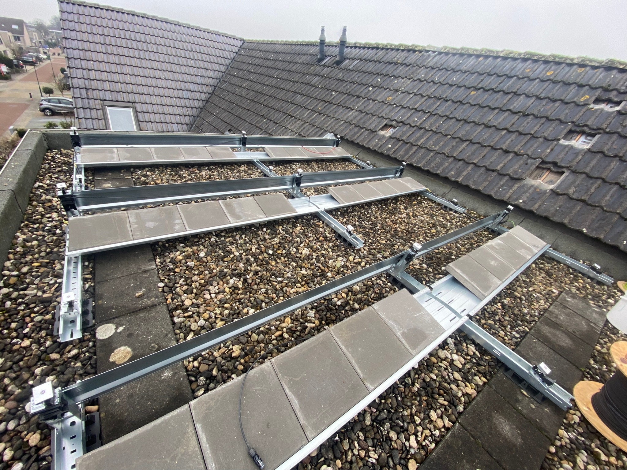 Montaż stelaża pod panele słoneczne na dachu pokrytym dachówką, widoczne elementy mocujące i kamyki na dachu.