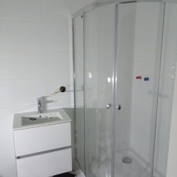 Łukrem - Nowoczesna łazienka z białą umywalką z szafką, szklaną kabiną prysznicową z szaro-białą mozaiką wewnątrz i czarno-białą szachownicą na podłodze, widoczny niedokończony montaż baterii.