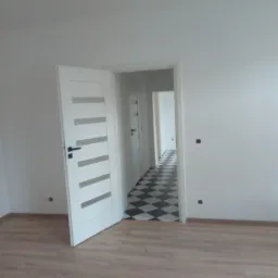 Wnętrze mieszkania po remoncie: widok na pokój z panelami podłogowymi, otwarte białe drzwi z przeszkleniami prowadzą do korytarza z czarno-białą podłogą w szachownicę, ściany pomalowane na biało.