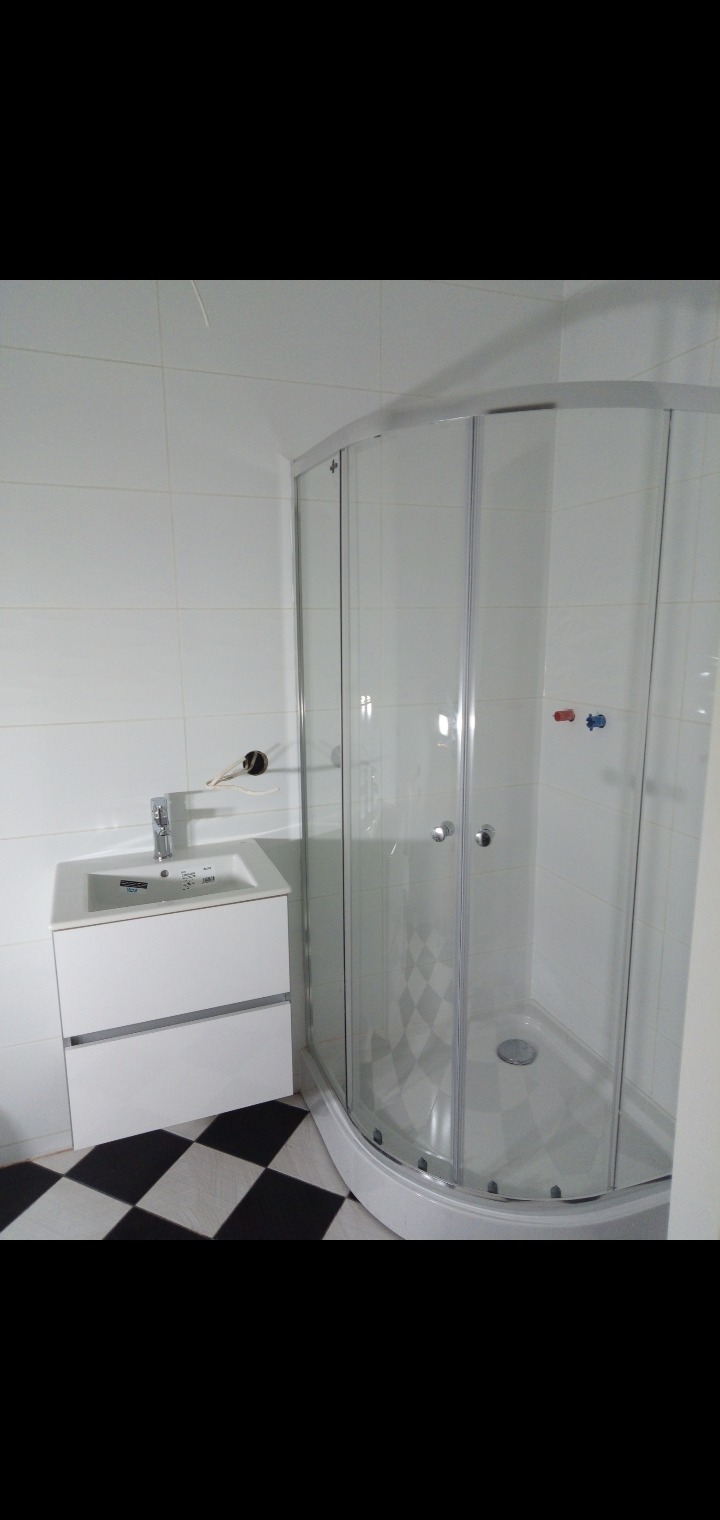 Nowoczesna łazienka z białą umywalką z szafką, szklaną kabiną prysznicową z szaro-białą mozaiką wewnątrz i czarno-białą szachownicą na podłodze, widoczny niedokończony montaż baterii.