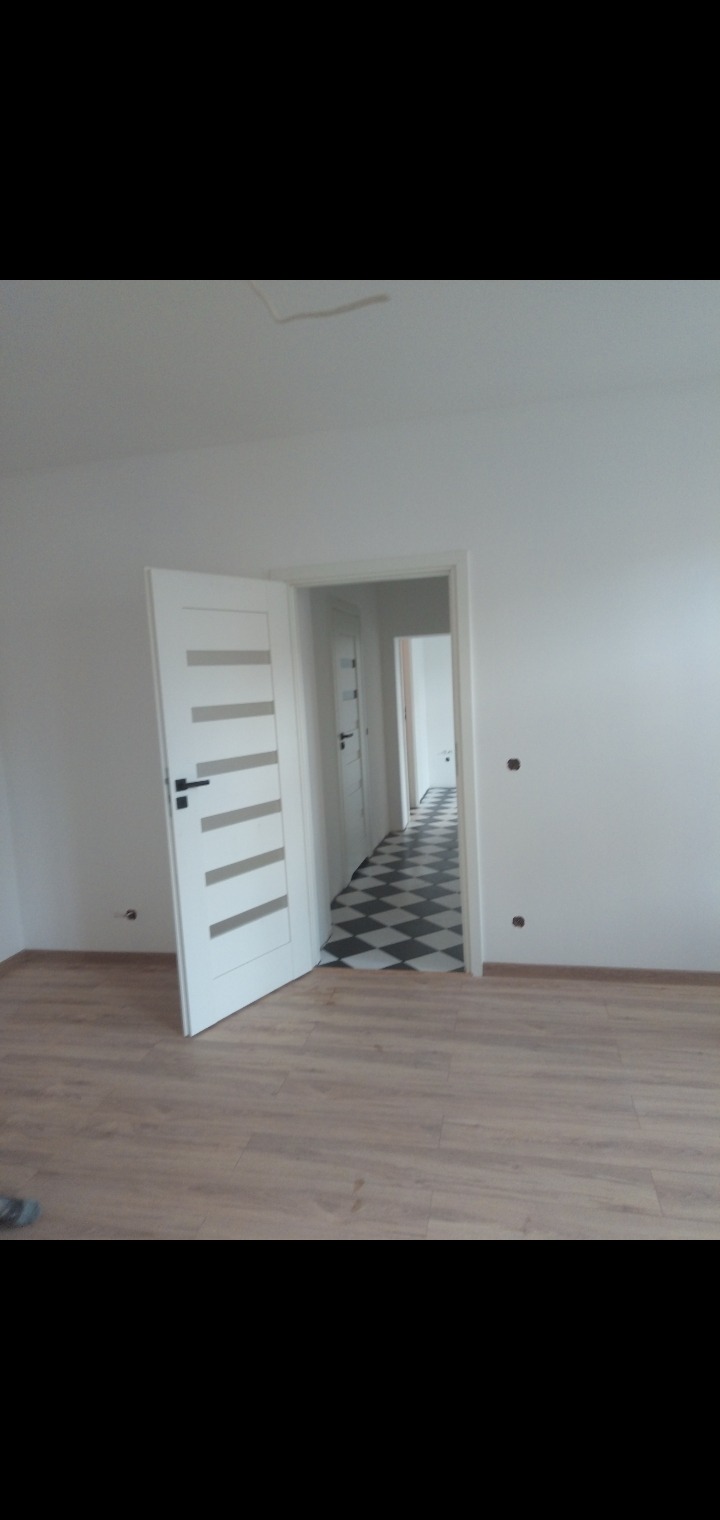 Wnętrze mieszkania po remoncie: widok na pokój z panelami podłogowymi, otwarte białe drzwi z przeszkleniami prowadzą do korytarza z czarno-białą podłogą w szachownicę, ściany pomalowane na biało.