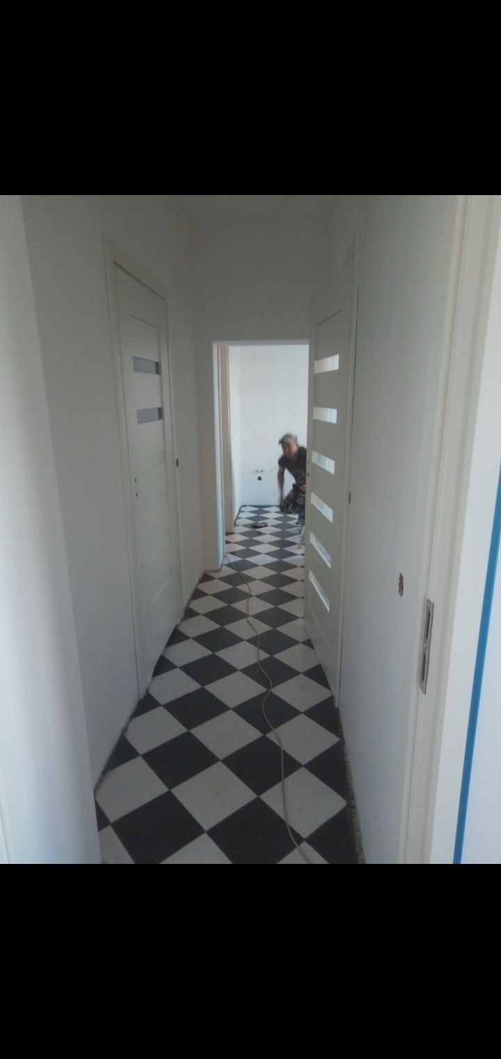 Korytarz z czarno-białą podłogą w romby, białe ściany i drzwi, w tle widoczny pracownik w trakcie układania podłogi w kolejnym pomieszczeniu.