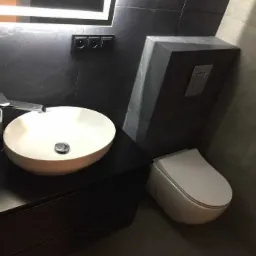 Nowoczesna łazienka w ciemnej tonacji z owalną umywalką nablatową, chromowaną baterią, podświetlanym lustrem i podtynkową toaletą.